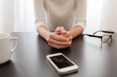 A woman sits alone at a table with her glasses off, her cell phone blank on the table in front of her, and an empty coffee mug to the side. 