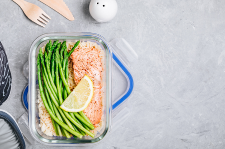 An aerial view of a glass container containing salmon and asparagus garnished with lemon.  The lid sits just under the container and nearby are a wooden knife, fork, and salt and pepper shaker