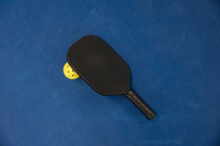 A black pickleball paddle and bright yellow ball resting on a dark blue court. 